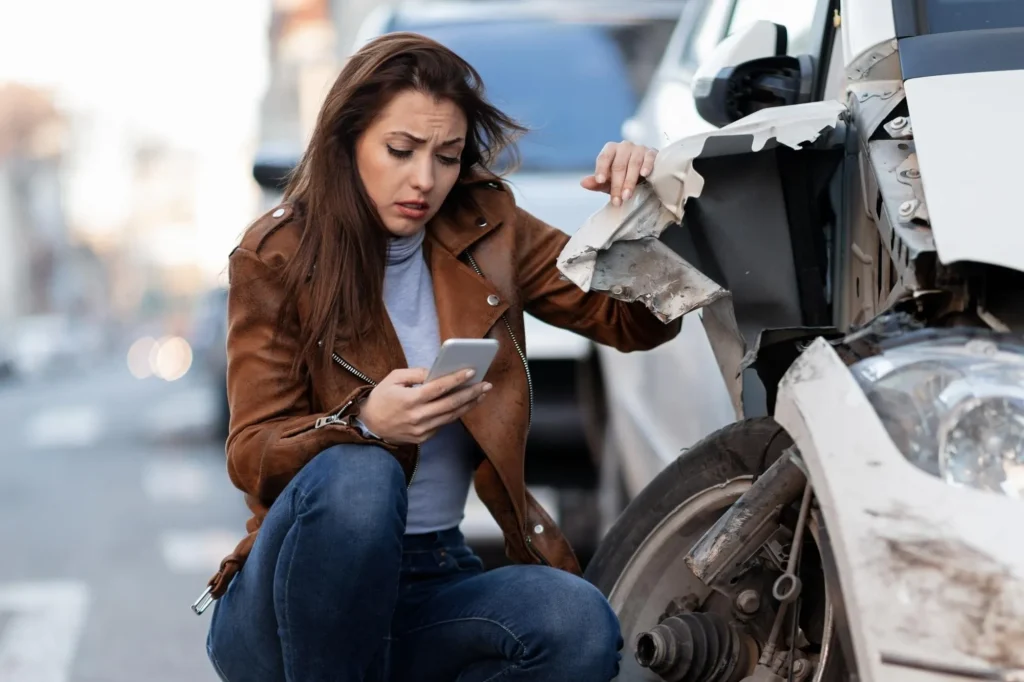 Woman in brown jacket using phone beside damaged white car on street.