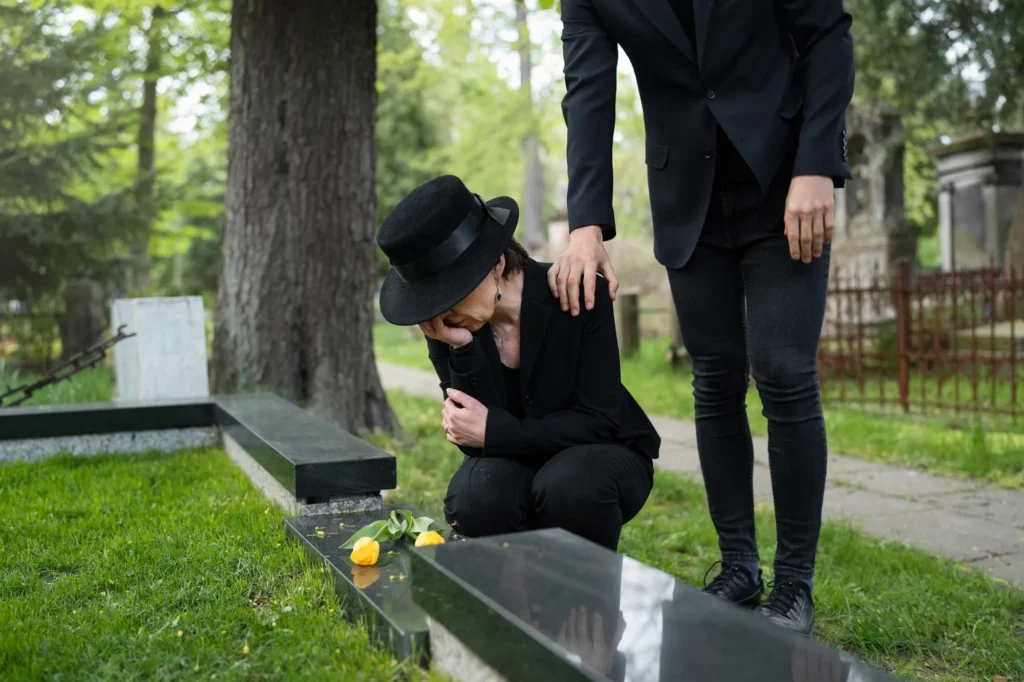 Woman in black mourning attire kneeling at grave with yellow flowers, comforted by standing person.