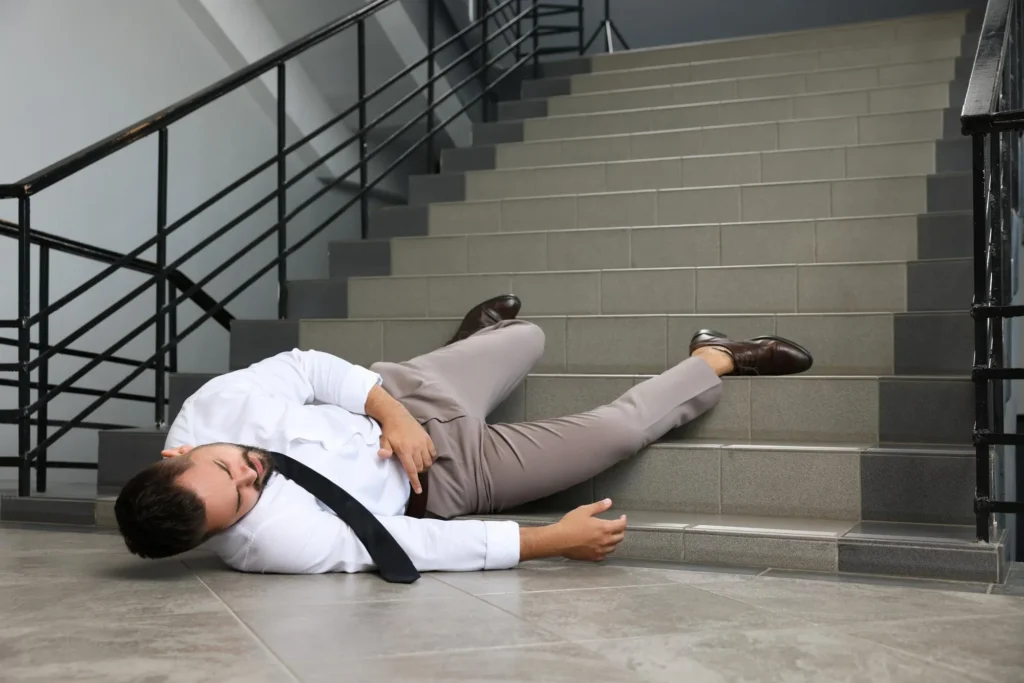 Man in business attire lying on tiled floor at bottom of indoor staircase, holding his side.
