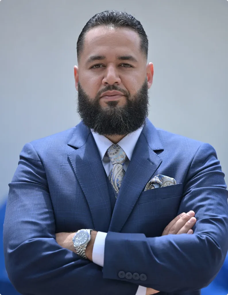Professional man with beard in blue suit, patterned tie, and silver watch, arms crossed.