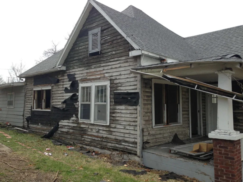 Exterior view of a weathered, damaged wooden house with peeling black siding and broken windows.