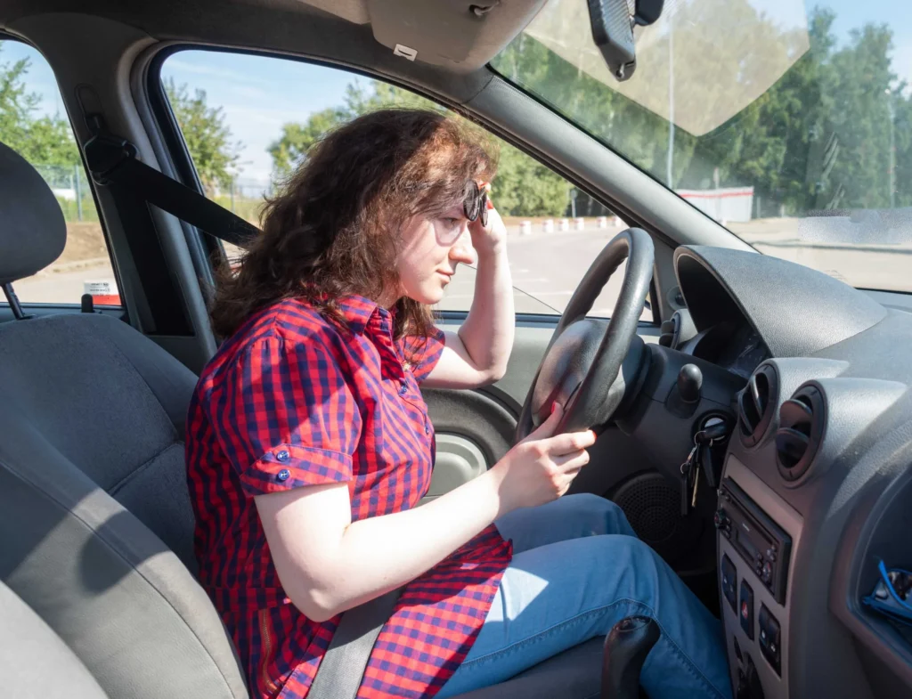 Woman in red and blue checkered shirt sitting in car driver's seat, holding steering wheel.