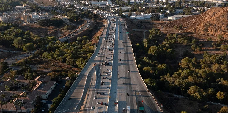 Aerial view of a multi-lane highway with moderate traffic surrounded by trees and residential areas.