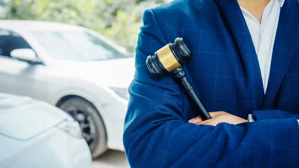 Person in blue suit holding a black and gold judge's gavel with crossed arms, near white cars.