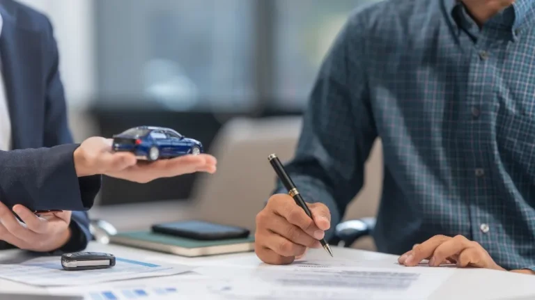 Two people at a desk, one holding a blue toy car, the other signing documents.