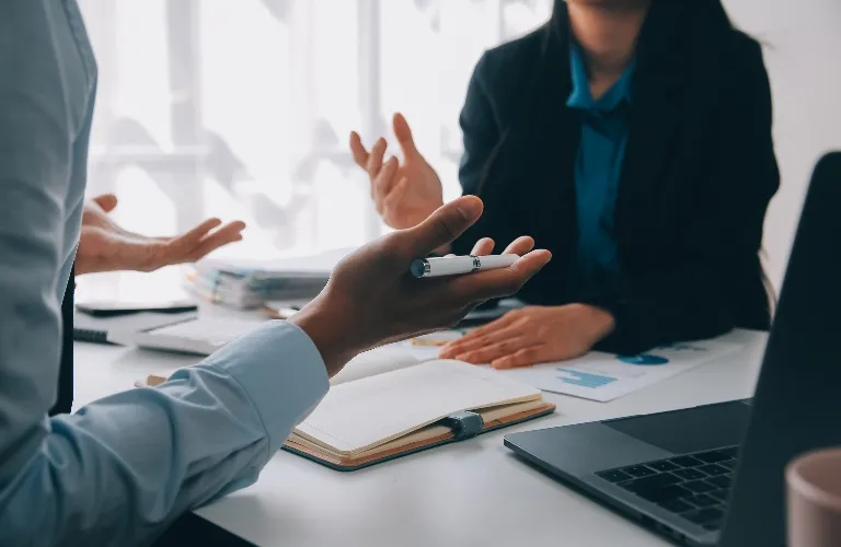 Two professionals engaged in discussion at a desk with documents and a laptop.