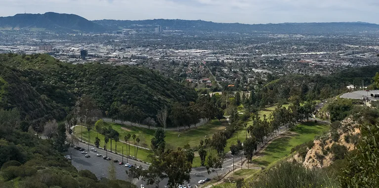 Panoramic view of a green park and parking lot surrounded by hills, overlooking a sprawling cityscape.