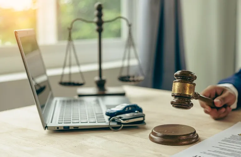 Wooden judge's gavel striking block beside laptop, car keys, and scales of justice.