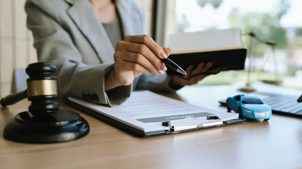 Person in gray blazer holding pen and notebook, with legal documents, gavel, and toy car on wooden desk.