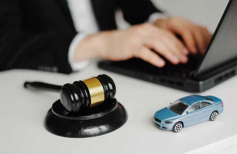 Black and gold judge's gavel and blue toy car on desk, person typing on laptop in background.