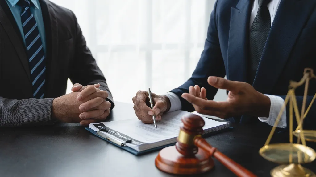 Two men in suits discussing legal documents at a desk with gavel and scales.