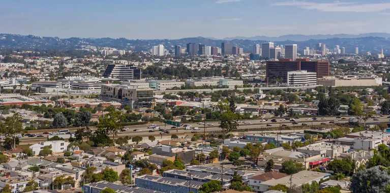Aerial view of urban cityscape with residential houses, highways, and distant high-rise buildings under a clear sky.