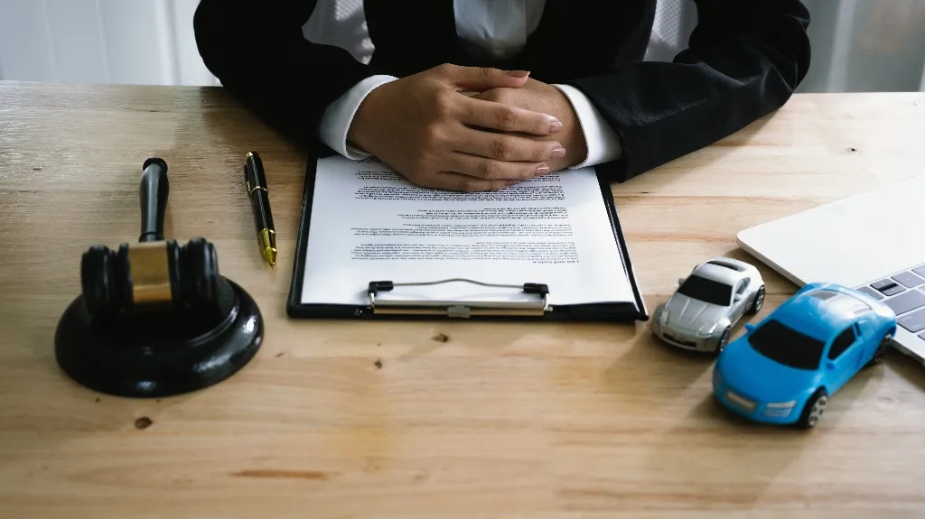 Person in formal attire seated at desk with legal document, gavel, pen, and toy cars.