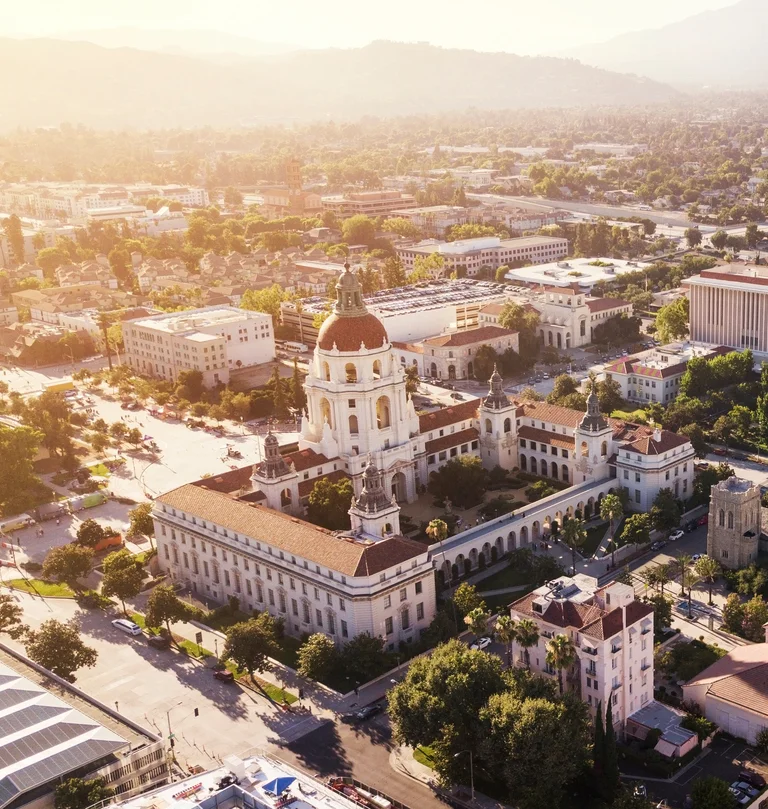 Aerial view of a historic building with a central dome and surrounding towers, situated in a sunny urban area with trees and mountains in the background.