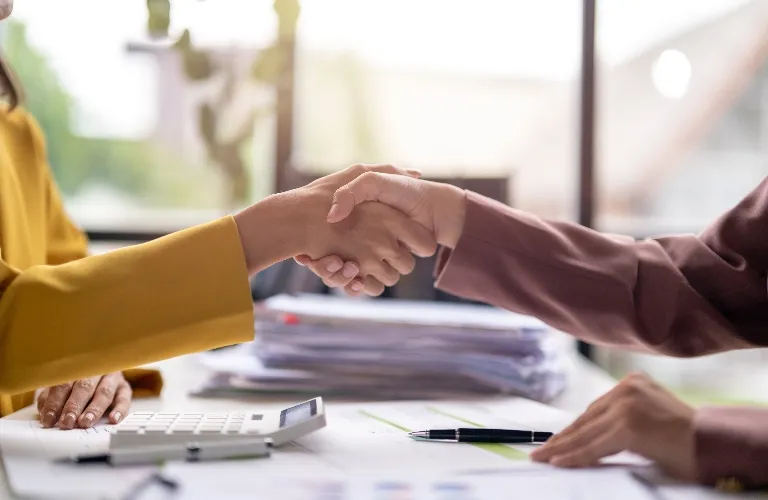 Two people shaking hands over a desk with documents, a pen, and a calculator.