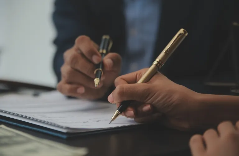 Close-up of two people holding pens over documents on a dark table.