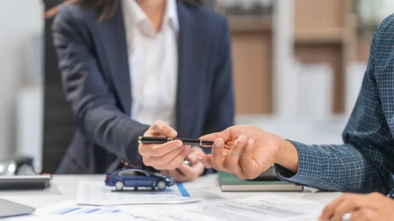 Two professionals exchanging a pen over a desk with documents and a blue toy car.