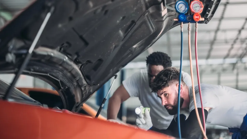 Two male mechanics in white shirts inspecting an orange car engine under the hood.