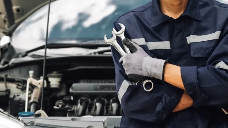 Mechanic in navy uniform holding wrenches with car engine visible in the background.