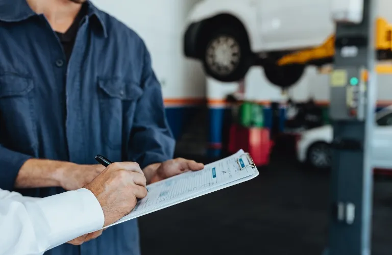 Two individuals in a garage, one writing on a clipboard, with a white car elevated on a hydraulic lift in the background.