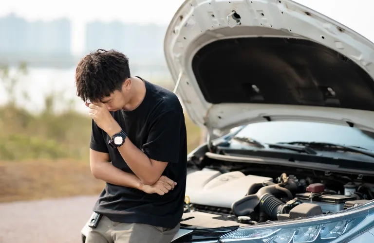 Young man in black shirt leaning against a white car with an open hood, outdoors.