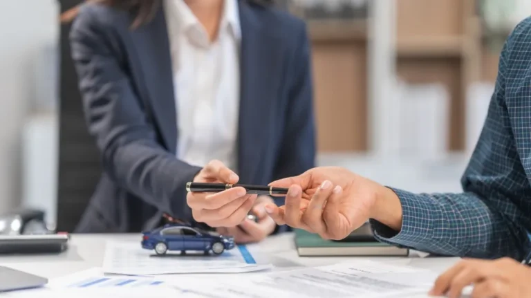Business professional in suit handing pen to client across desk with car model and documents.
