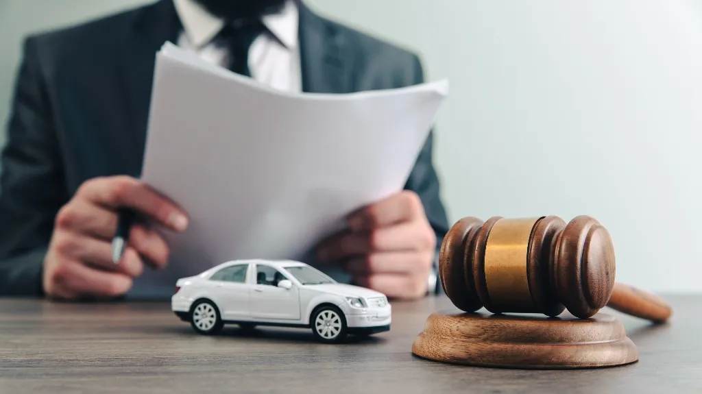 Man in suit holding documents, with wooden judge's gavel and white toy car on table.