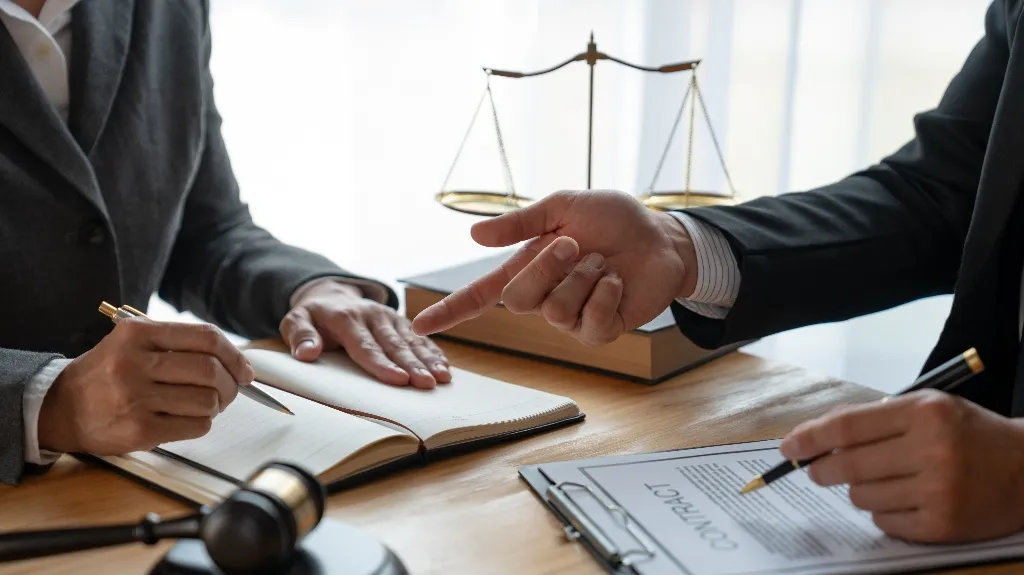 Two professionals in suits reviewing a contract with legal scales and a gavel on a wooden desk.