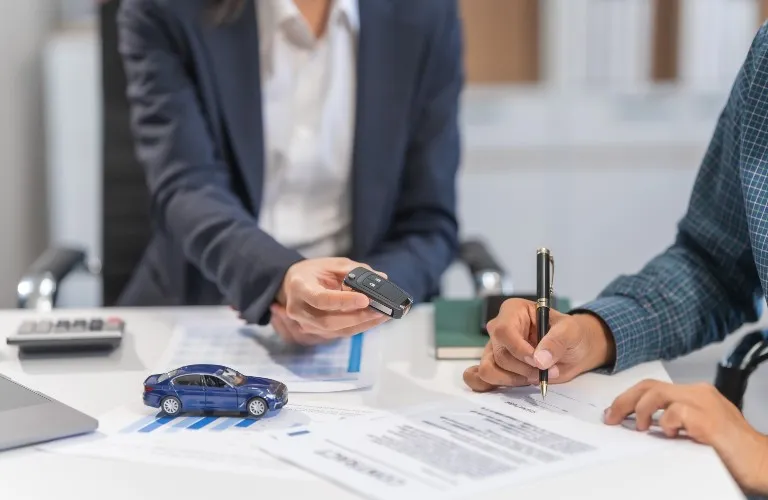 Two individuals at a desk exchanging car keys and signing documents, with a blue toy car on the table.