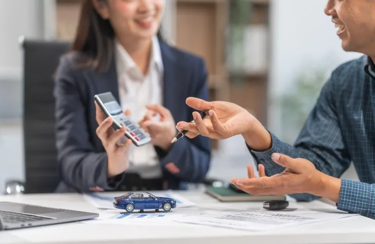Two people discussing car sale with miniature blue car, car keys, and calculator on table.
