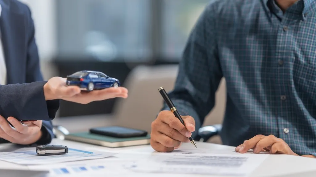 Two individuals at a desk, one holding a blue toy car and the other signing documents with a pen.