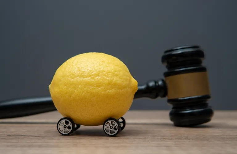 A yellow lemon with small car wheels attached, positioned in front of a wooden judge's gavel on a wooden surface.