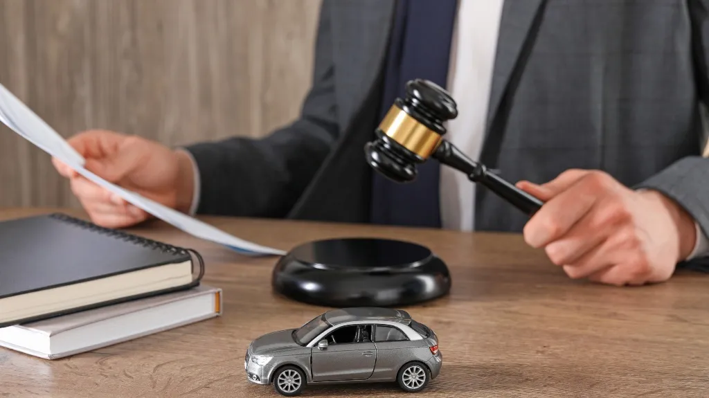Person in suit holding gavel and document, with toy car and books on wooden desk.
