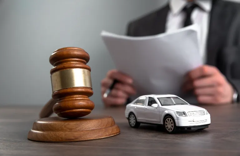 Wooden judge's gavel and silver toy car on table, person in suit reviewing documents in background.
