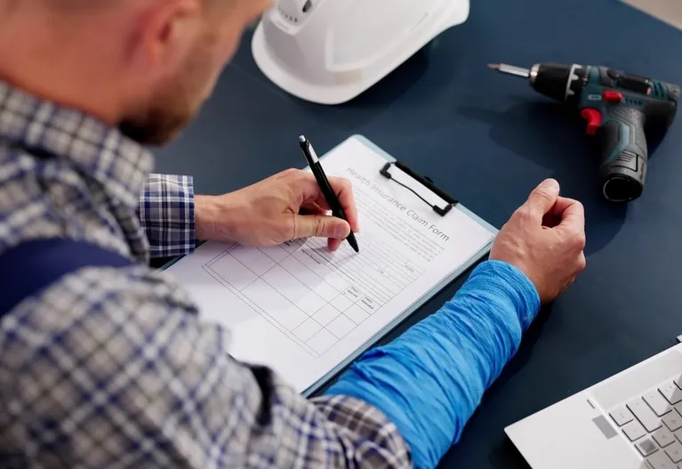 Man in plaid shirt completing health insurance claim form on clipboard, with white hard hat and electric drill on desk.