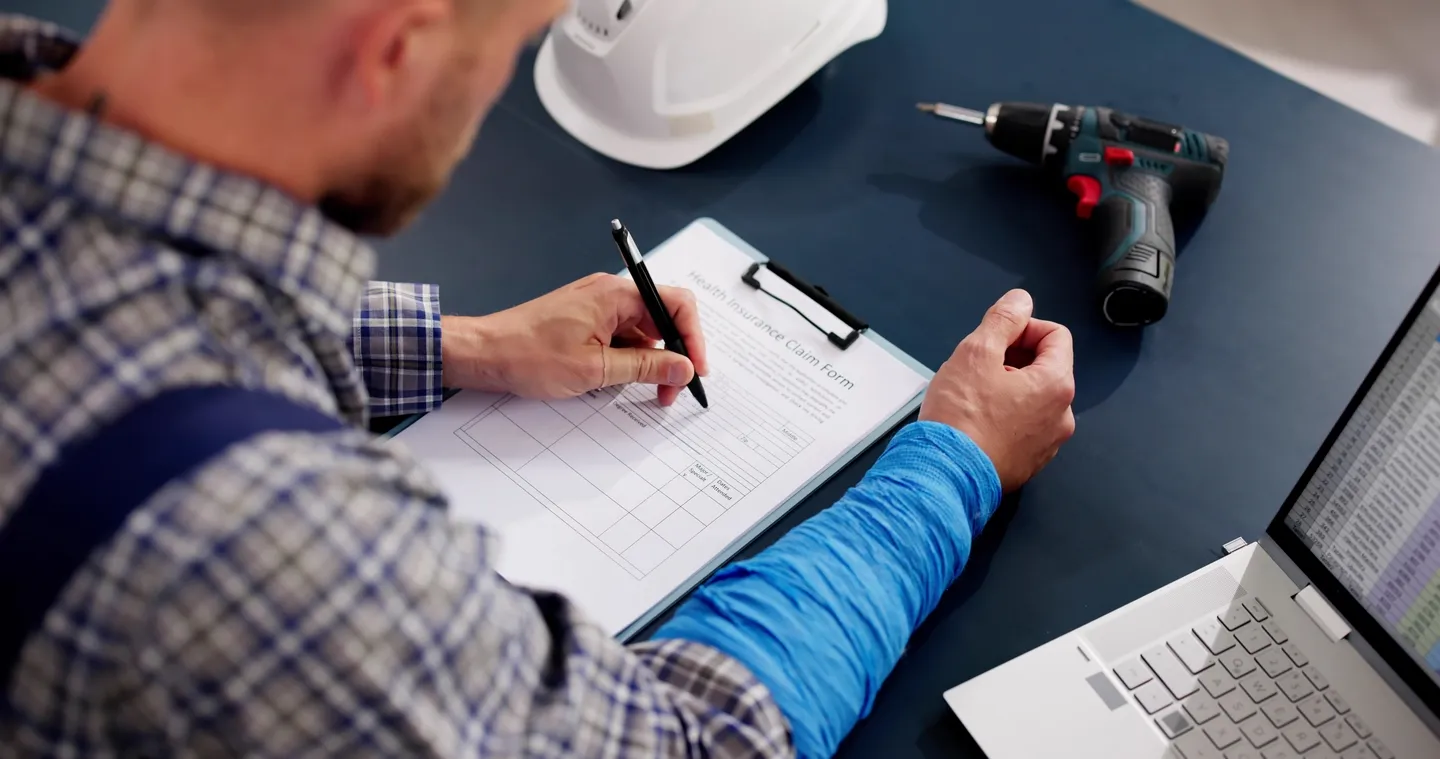Man in plaid shirt filling out health insurance claim form, with hard hat, drill, and laptop on desk.