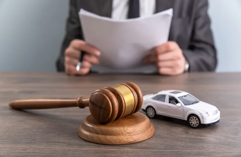Wooden judge's gavel and toy white car on desk with person in suit holding documents.