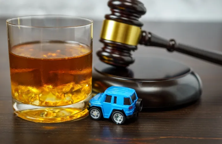 Glass of whiskey with ice, blue toy car, and wooden judge's gavel on table.