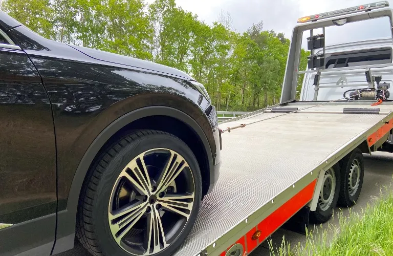 Black vehicle being loaded onto a silver flatbed tow truck on a roadside.