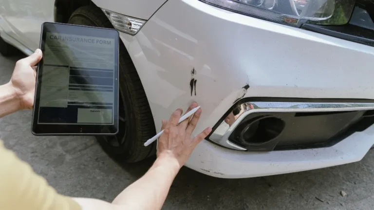 Person holding a tablet with a car insurance form, inspecting damage on white car bumper.