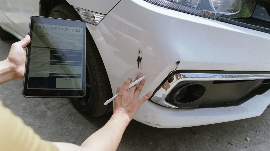 Person holding a tablet with a car insurance form, inspecting damage on white car bumper.