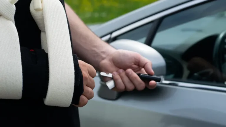 Person with arm in white sling holding a car key near a silver vehicle door.