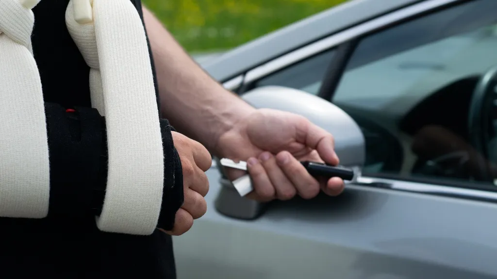Person with arm in white sling holding a car key near a silver vehicle door.