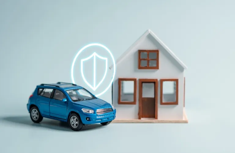 Blue toy car and small white model house with glowing shield icon, symbolizing insurance.