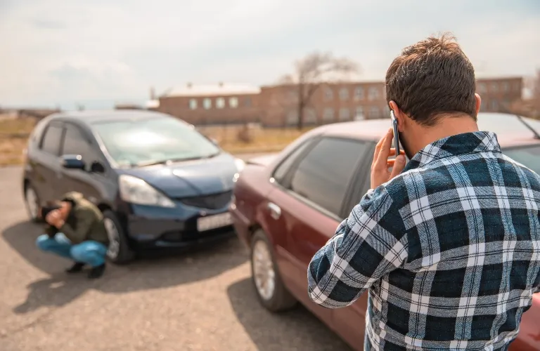 Man in plaid shirt making a phone call near two cars involved in a minor accident.