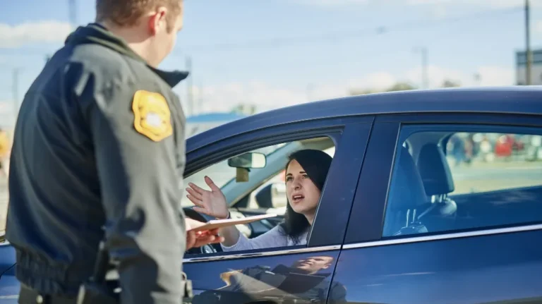 Police officer in black uniform speaking to woman driver in blue car during traffic stop.
