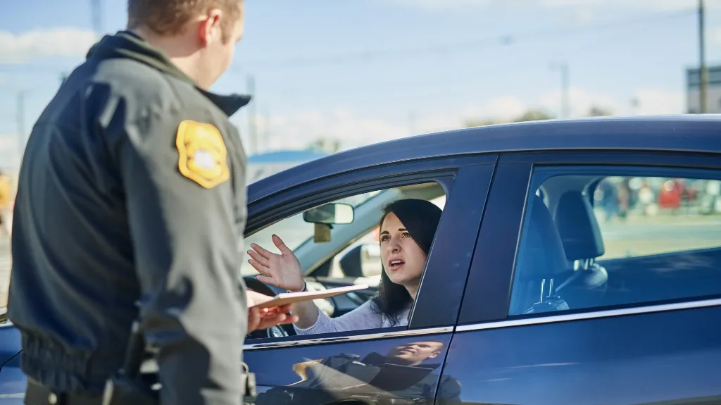 Police officer in black uniform speaking to woman driver in blue car during traffic stop.