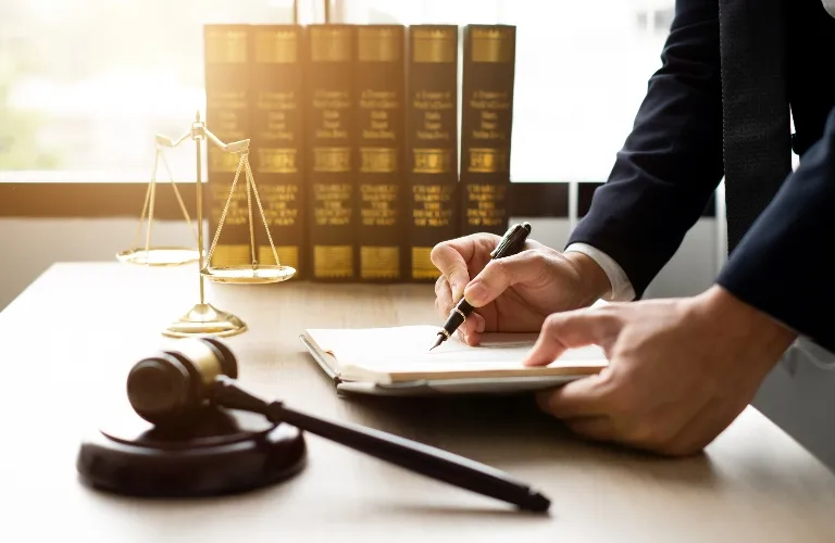 Person in business attire writing on a notepad at a desk with legal books, scales of justice, and a gavel.